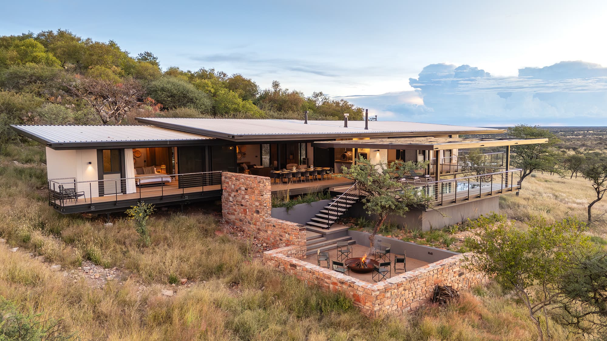 Modern luxury lodge featuring a stone fire pit and deck overlooking a vast savanna landscape.