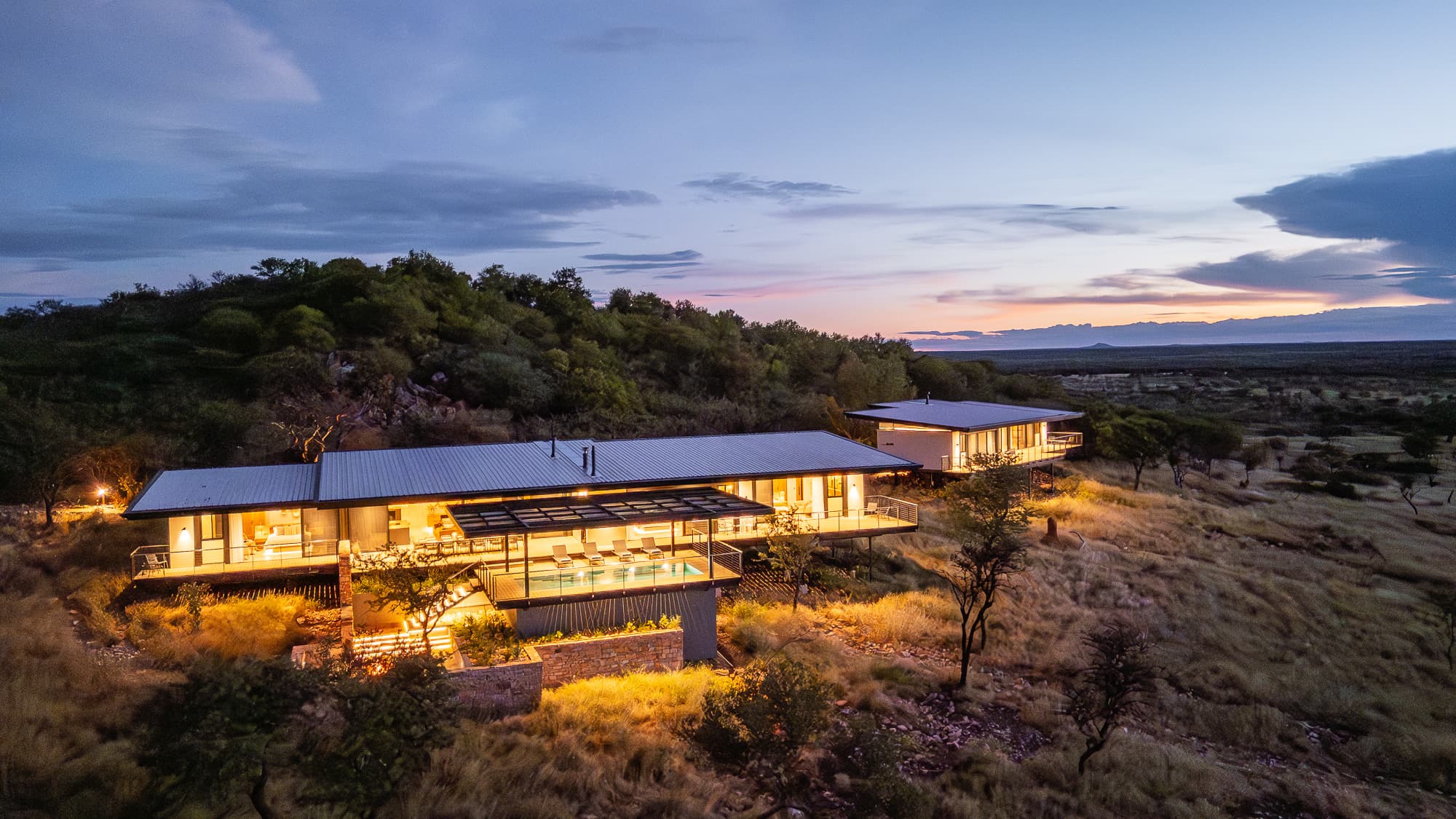 Modern hillside villa with glowing lights and a pool deck overlooking a vast savanna.