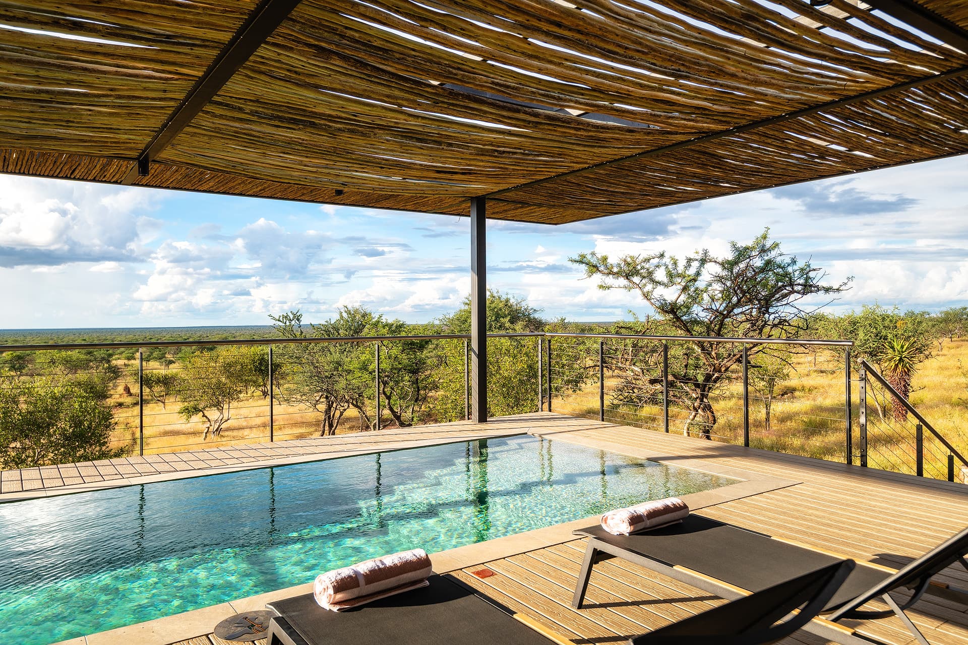 Luxury pool deck with lounge chairs under a stick pergola overlooking a scenic savanna.
