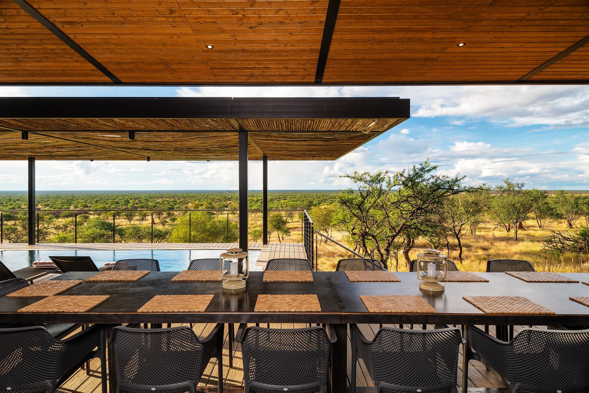Long dining table with lanterns overlooking a pool and expansive savanna landscape from a deck.