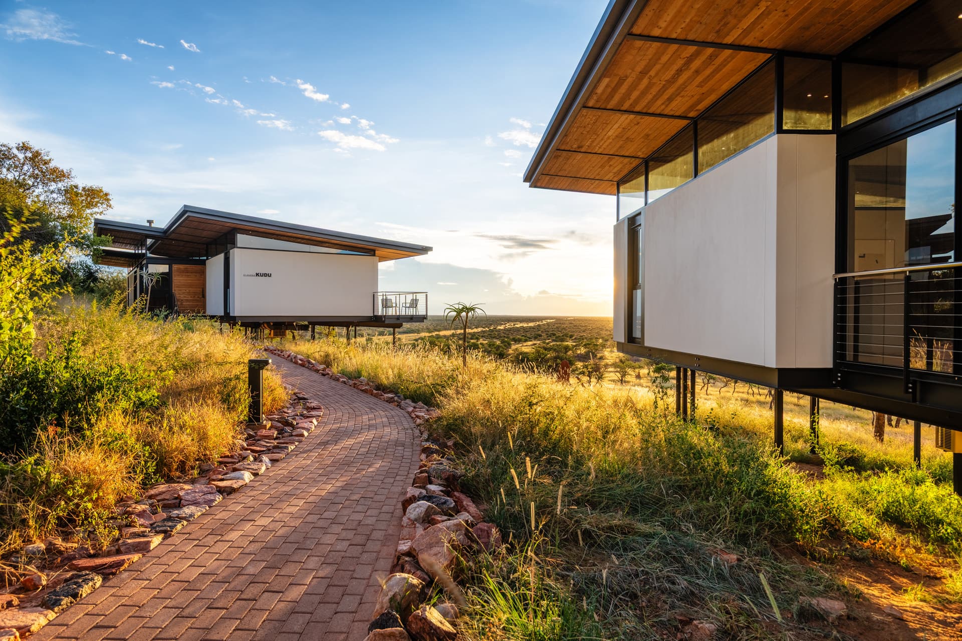 Modern elevated lodges overlooking a golden savanna at sunset with a winding brick-paved path.