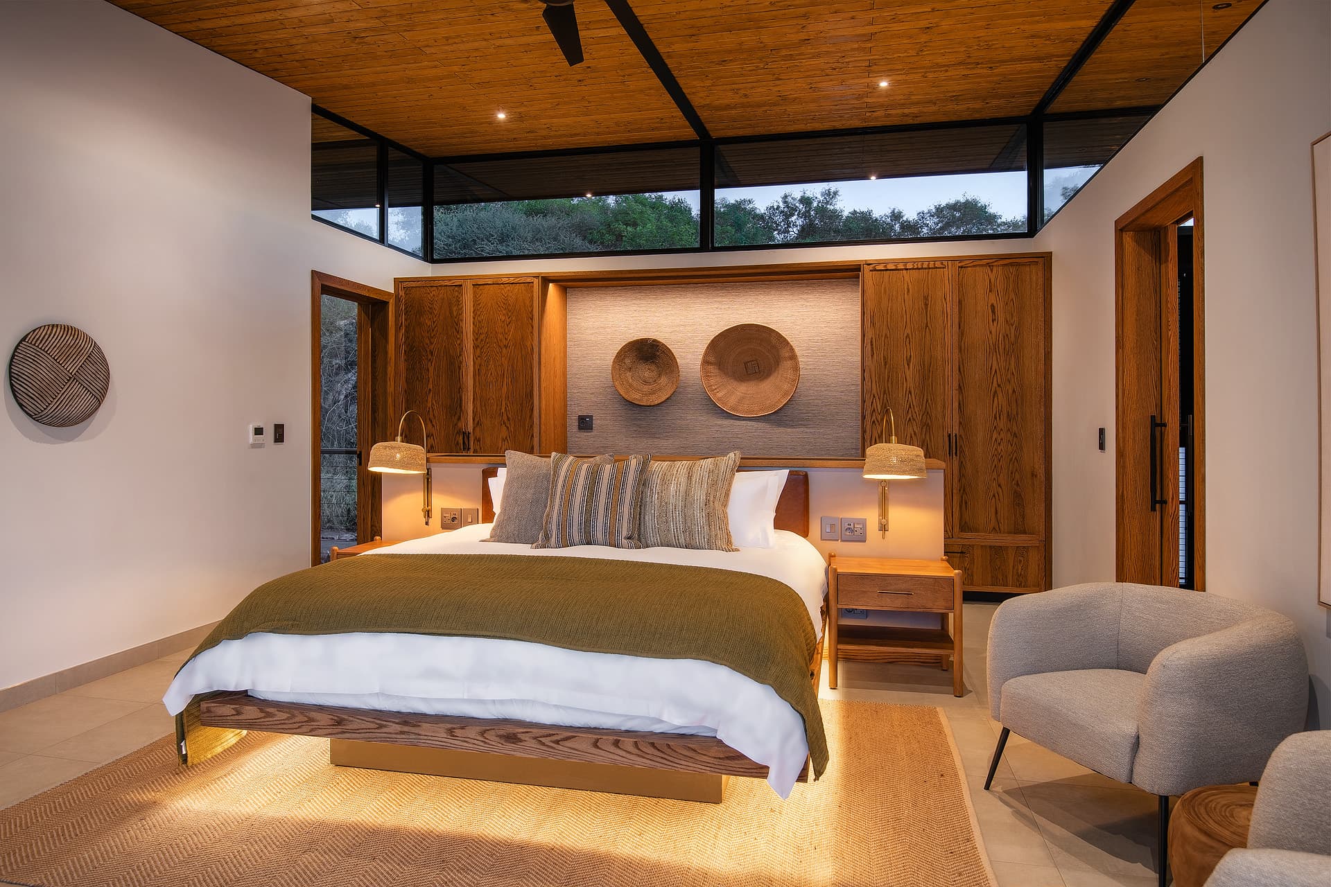 Modern bedroom with wooden ceiling, woven wall decor, warm lighting, and windows overlooking trees.