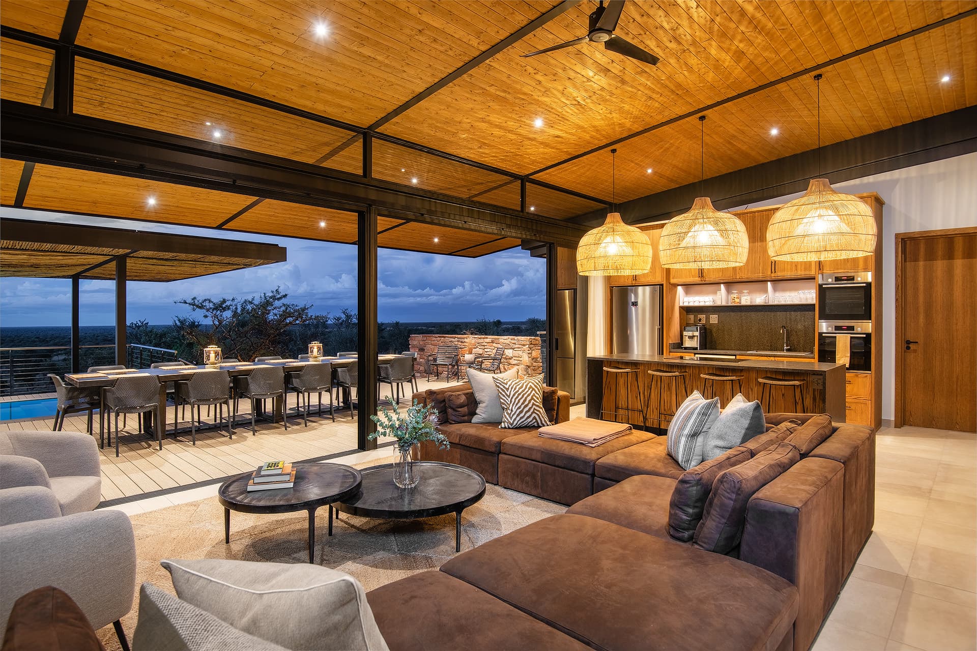 Modern open-plan living room with a brown sectional sofa and kitchen overlooking a scenic deck.