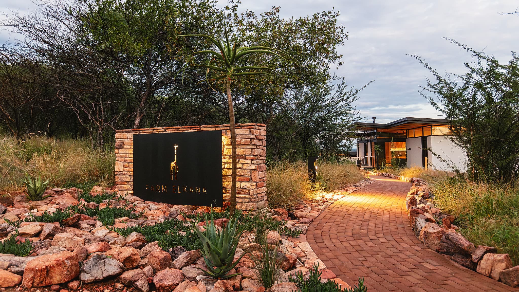 Illuminated Farm Elkana sign and brick path leading to a modern lodge among trees.