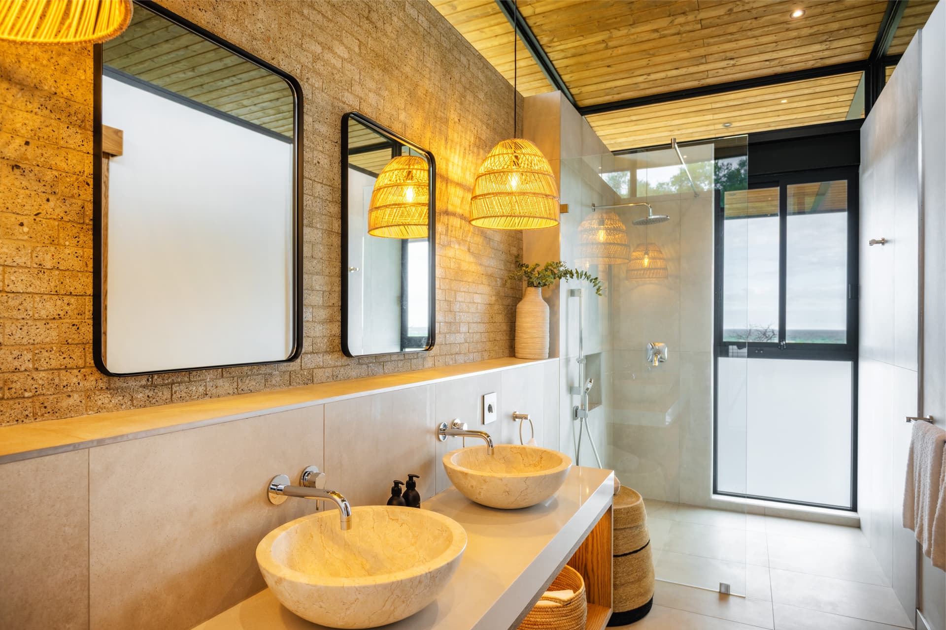 Modern bathroom with stone vessel sinks, woven lights, brick walls, and a glass-enclosed shower.